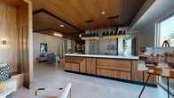 This thoughtfully designed single-floor amenity space blends mid-century modern aesthetics with contemporary finishes to create a welcoming and visually dynamic environment. The centerpiece is a striking bar and reception counter crafted from richly grained wood veneer, topped with a crisp white surface and framed by a decorative geometric lattice partition. An eye-catching pendant light fixture with black angular shades and gold fringe accents hangs overhead, adding a bold focal point to the space.

Adjacent to the bar, a dining area provides ample seating at white-topped tables with upholstered grey and gold chairs, set against textured herringbone tile accent walls and flanked by sculptural art pieces and vibrant yellow figurative artwork. A beverage station is conveniently positioned nearby.

The lounge areas feature plush seating arrangements with tan leather chairs, cushioned sofas set into warm wood-paneled nooks, round mirrors, and lush tropical plants that bring life and greenery throughout. Large glass doors open to an outdoor terrace, flooding the interior with natural light and offering views of the surrounding area.

A corridor leads to a fully equipped fitness center, framed by bold chevron-patterned dark accent walls and glass-panel doors. Throughout the space, large-format light tile flooring, wood-panel ceilings, and curated contemporary artwork contribute to a cohesive, sophisticated ambiance.