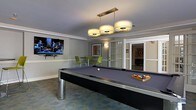 This well-appointed single-floor amenity space is designed for both recreation and productivity. The centerpiece of the main room is a full-size pool table with a slate-blue felt surface, set beneath a modern three-drum pendant light fixture with brushed brass accents that provides both style and focused illumination. Crown molding runs along the perimeter of the ceiling, adding a polished, refined touch to the overall aesthetic. A large flat-screen television is mounted on the wall, making the space equally suited for casual gatherings and entertainment. Adjacent to the billiards area, a small lounge nook features a high-top table with vibrant green chairs, offering a spot for socializing or casual dining. The flooring features a patterned carpet that adds texture and visual interest throughout. Glass-paneled French doors lead to a connected room, identified by a prominent "@" symbol signage, suggesting a dedicated business center or computer lounge. The walls are finished in neutral, warm tones that create an inviting and comfortable atmosphere. Overall, the space strikes a balance between leisure and functionality, making it a versatile amenity suitable for a variety of uses and occasions.