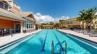 Located at 9505 Blackwood Circle in Fort Myers, Florida, this outdoor amenity area offers a well-appointed resort-style pool complex designed for both recreation and relaxation. The centerpiece is a generously sized lap pool with clearly marked lanes, ideal for fitness swimming, complemented by a separate leisure pool area equipped with a decorative fountain feature. The pool deck is finished with attractive paver stonework and is lined with lounge chairs and seating areas, providing ample space for sunbathing and socializing. A charming clubhouse structure anchors the space, featuring a warm terracotta and earth-toned exterior with a tile roof, large windows, and a covered porch with a pergola-style overhang. Outdoor dining tables and umbrellas offer shaded gathering spots nearby. The surrounding landscape is lush with mature palm trees and manicured hedging, creating a private, tropical atmosphere. The community buildings visible beyond the pool area reflect a cohesive architectural palette of warm yellows, oranges, and reds with tile roofing. The entire outdoor space benefits from the abundant natural light and clear blue skies characteristic of Southwest Florida, making it an inviting year-round retreat for residents.