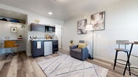 This efficiently designed single-floor studio offers a thoughtfully arranged open-plan layout that maximizes every square foot. The main living area features light wood-style vinyl flooring throughout, crisp white walls, and crown molding details that lend a clean, polished aesthetic. A wall-mounted flat-screen TV anchors one side of the room, while a geometric-patterned area rug defines the lounge space, complemented by a convertible sofa chair — ideal for flexible living.

The kitchenette is smartly appointed with dark wood-finish cabinetry, an under-counter refrigerator, a built-in microwave, a single-basin sink with a checkered tile backsplash, and open shelving for storage. A compact bistro-style high-top table with two stools sits beside a window with slatted blinds, creating a charming dining nook bathed in natural light.

The private bathroom continues the wood-style flooring and features a shower with a curtain enclosure, a toilet, and a window for ventilation and natural light. The overall ambiance of the space is bright, modern, and functional — making it a practical and inviting self-contained living unit.