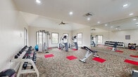 This fitness center in Cordova, Tennessee, is a single-level facility offering a thoughtfully organized workout environment. The space features vaulted ceilings that lend an open, airy feel, complemented by recessed lighting and a ceiling fan that ensure comfortable conditions during exercise. Patterned carpet tiles with red accents cover the flooring throughout, while red exercise mats are strategically placed beneath equipment stations.

The facility is divided into distinct workout zones. The cardio area houses multiple treadmills, elliptical trainers, and a stair climber, each positioned in front of large windows that overlook the outdoor surroundings, including a pool area. Wall-mounted flat-screen televisions are positioned above the cardio machines, providing entertainment during workouts. The strength training zone includes a multi-station cable machine, a dedicated dumbbell rack with a full range of weights, a stability ball, and an adjustable weight bench.

Large mirrors line one full wall, visually expanding the space and allowing users to monitor their form. Multiple windows distributed across the walls flood the interior with natural light. The neutral-toned walls and clean, organized layout contribute to a bright, functional, and motivating atmosphere suited for a variety of fitness routines.