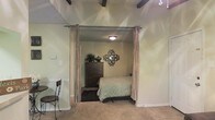 This single-story space showcases a thoughtfully arranged interior with distinct character. The most notable architectural feature is the exposed dark ceiling beams overhead, which add a rustic, structural element to the overall design. The bedroom area is defined by floor-to-ceiling curtain panels mounted on a rod, creating a soft, elegant room divider that allows for flexible separation of spaces while maintaining an open feel.

Inside the sleeping area, neutral-toned walls provide a calm backdrop, complemented by a flush-mount ceiling light that offers warm, even illumination. A dark wood dresser serves as a functional storage piece, topped with a decorative vase and flanked by a lush potted plant that adds a natural element. An ornate metal wall mirror with a geometric design acts as a decorative focal point above the dresser.

The bedroom features carpeted flooring, while the adjacent area transitions to tile, distinguishing the two zones. A bistro-style chair and small table are visible in the foreground, suggesting a casual seating nook. The overall ambiance is warm and homey, with layered textures and decorative touches contributing to a comfortable, lived-in aesthetic.