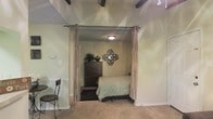 This single-story space showcases a thoughtfully arranged interior with distinct character. The most notable architectural feature is the exposed dark ceiling beams overhead, which add a rustic, structural element to the overall design. The bedroom area is defined by floor-to-ceiling curtain panels mounted on a rod, creating a soft, elegant room divider that allows for flexible separation of spaces while maintaining an open feel.

Inside the sleeping area, neutral-toned walls provide a calm backdrop, complemented by a flush-mount ceiling light that offers warm, even illumination. A dark wood dresser serves as a functional storage piece, topped with a decorative vase and flanked by a lush potted plant that adds a natural element. An ornate metal wall mirror with a geometric design acts as a decorative focal point above the dresser.

The bedroom features carpeted flooring, while the adjacent area transitions to tile, distinguishing the two zones. A bistro-style chair and small table are visible in the foreground, suggesting a casual seating nook. The overall ambiance is warm and homey, with layered textures and decorative touches contributing to a comfortable, lived-in aesthetic.