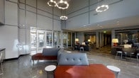 This single-floor lobby presents a sophisticated and contemporary design, characterized by dramatic double-height ceilings that lend an impressive sense of openness and grandeur. A series of circular pendant light fixtures with polished metallic finishes hang at varying heights, creating a striking focal point overhead while providing warm, ambient illumination throughout the space.

The seating area features a curated arrangement of tufted gray sofas and bold rust-orange benches, offering a modern contrast in both texture and color. Small accent tables complement the lounge area, which is anchored by a large area rug beneath a central concierge or reception desk, complete with task lighting and office seating for staff.

Floor-to-ceiling glass walls and doors along the entry facade flood the interior with abundant natural light, providing clear sightlines to an exterior courtyard or outdoor area beyond. The flooring consists of large-format stone or tile in a neutral gray tone, reinforcing the polished, professional aesthetic.

To the rear of the lobby, corridors lead deeper into the building, with additional architectural columns and accent lighting maintaining the refined atmosphere throughout. The overall ambiance is sleek, welcoming, and distinctly upscale.