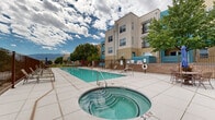 This outdoor amenity area centers around a generously sized rectangular swimming pool with clear turquoise water, designed for both leisure and lap swimming. Adjacent to the pool, a circular hot tub provides a dedicated space for relaxation. The surrounding deck is finished in large-format light-toned tile, offering ample space for sunbathing and socializing, and is furnished with an array of lounge chairs and patio seating sets, including umbrella-shaded tables ideal for outdoor dining or casual gatherings. The entire area is enclosed by a wrought iron fence, providing both security and an open, airy feel that allows unobstructed views of the surrounding desert landscape and distant mountain ranges. Mature trees are situated along the perimeter, offering natural shade and greenery that contrast with the arid surroundings. The multi-story residential building adjacent to the pool area features a contemporary design with a neutral stucco exterior accented by bold teal and blue panels, along with private balconies that overlook the pool deck. The pool area benefits from abundant natural sunlight under expansive southwestern skies, creating a bright and inviting atmosphere throughout the day. Safety equipment, including a life ring, is visibly posted on-site.