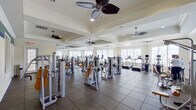 This fitness center offers a comprehensive, single-floor workout facility designed to accommodate a wide range of exercise needs. The main workout floor is generously sized, featuring tray ceilings with multiple ceiling fans that promote air circulation and comfort throughout. Large windows line the walls, flooding the space with abundant natural light and providing views of the surrounding exterior. Full-length mirrors are positioned along one wall, enhancing the sense of openness and allowing users to monitor their form during exercise.

The main floor is outfitted with a robust selection of professional-grade Cybex strength training machines, featuring padded yellow and tan seating, as well as multiple treadmills and cardio equipment arranged along the windowed wall. The flooring combines large-format tile with rubberized mat sections beneath the equipment, ensuring durability and safety.

An adjacent, glass-windowed room serves as a dedicated free weights area, visible from the main floor and accessible through French doors. This secondary space provides a more intimate setting for dumbbell and targeted strength work. A wall-mounted flat-screen television is also present, adding an entertainment option for users. The overall ambiance is clean, bright, and functional, reflecting a well-maintained facility built for serious fitness use.
