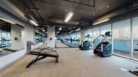 Located at 1000 S Clark Street in Chicago's South Loop neighborhood, this fitness center offers a comprehensive, well-equipped workout environment spread across a single expansive floor. The facility features an industrial-modern aesthetic, with exposed ductwork and structural elements painted in dark tones against light walls, complemented by bright linear lighting throughout.

The cardio zone lines the perimeter, where floor-to-ceiling glass windows provide sweeping views of the Chicago skyline and surrounding greenery — an energizing backdrop for treadmill runs, elliptical sessions, stair climbers, rowing machines, and stationary bikes. Large wall mirrors amplify the sense of space and allow users to monitor their form across all areas.

A dedicated strength and free weights section offers adjustable benches, cable machines, and a variety of resistance equipment. Rubber flooring in the strength zones provides durability and safety, while carpet tiles cover the cardio and open stretching areas for comfort underfoot.

An open functional training area provides ample space for dynamic movement, stretching, and floor-based exercises. A mounted flat-screen television and water fountain add practical amenities. The layout flows intuitively between zones, making the facility accessible and efficient for all fitness levels.