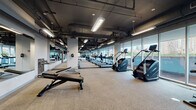Located at 1000 S Clark Street in Chicago's South Loop neighborhood, this fitness center offers a comprehensive, well-equipped workout environment spread across a single expansive floor. The facility features an industrial-modern aesthetic, with exposed ductwork and structural elements painted in dark tones against light walls, complemented by bright linear lighting throughout.

The cardio zone lines the perimeter, where floor-to-ceiling glass windows provide sweeping views of the Chicago skyline and surrounding greenery — an energizing backdrop for treadmill runs, elliptical sessions, stair climbers, rowing machines, and stationary bikes. Large wall mirrors amplify the sense of space and allow users to monitor their form across all areas.

A dedicated strength and free weights section offers adjustable benches, cable machines, and a variety of resistance equipment. Rubber flooring in the strength zones provides durability and safety, while carpet tiles cover the cardio and open stretching areas for comfort underfoot.

An open functional training area provides ample space for dynamic movement, stretching, and floor-based exercises. A mounted flat-screen television and water fountain add practical amenities. The layout flows intuitively between zones, making the facility accessible and efficient for all fitness levels.