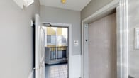 This single-floor interior hallway serves as a transitional common area within a larger multi-unit building. The space is finished with clean, light grey walls accented by white trim and crown molding, lending a crisp and well-maintained appearance throughout. Tile flooring in a neutral tone runs underfoot, contributing to the practical and durable character of the corridor.

A standout feature is the elevator access point, marked with a standard blue accessibility symbol, providing convenient vertical access within the building. Adjacent to the elevator, a white paneled door opens to further areas of the floor. At the end of the hallway, a doorway with a glazed panel opens onto a small outdoor balcony enclosed by a metal railing, offering a view of the surrounding building exterior, which features yellow and grey facades. Safety signage is posted near the balcony entrance, indicating fire safety awareness.

Overhead, a yellow ceiling fixture provides warm ambient lighting that complements the natural light filtering in from the balcony-facing window. The overall ambiance is functional and orderly, reflecting a well-kept residential or mixed-use building common area designed with both accessibility and everyday practicality in mind.