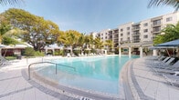This inviting outdoor amenity space centers around a generously sized resort-style swimming pool with crystal-clear turquoise water, designed with a gracefully curved perimeter and a gradual entry point marked at a shallow depth. The pool deck is finished with elegant patterned tile work and is lined with rows of white lounge chairs, offering ample space for relaxation under the open sky. Shaded pergola structures and cabanas are positioned along the perimeter, providing comfortable retreats from the sun. Tall, mature palm trees frame the area alongside established shade trees, lending a lush, tropical ambiance to the setting. A covered pavilion with a standing seam roof is visible at the far end, adding a functional architectural element to the outdoor environment. The backdrop features a well-appointed multi-story residential building with private balconies, contemporary white stucco facades, and large windows that overlook the pool area. The overall design evokes a warm, Florida-inspired aesthetic that seamlessly blends outdoor leisure with upscale residential living, making it an ideal gathering and recreation destination.
