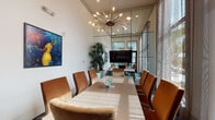 Located at 309 Mirroton Ave in Lakeland, Florida, this single-floor conference space delivers a polished and professional environment designed to impress. The room is anchored by a substantial wood conference table surrounded by plush cognac-upholstered chairs, comfortably accommodating groups for meetings or collaborative sessions. Soaring double-height ceilings lend a sense of grandeur, while dramatic floor-to-ceiling windows flood the interior with natural light and frame lush outdoor views of palm trees and the surrounding community. Sheer drapery softens the light and adds an elegant touch to the expansive glass facade. A striking sputnik-style brass chandelier serves as a bold focal point overhead, complementing the room's contemporary aesthetic. A large wall-mounted flat-screen television is positioned at one end of the room for presentations and media display, supported by a sleek credenza. A mirrored glass partition wall with black steel framing connects the conference room to an adjacent corridor, enhancing the sense of depth and openness. An artistic canvas featuring vibrant blue tones adds a creative, curated accent to the crisp white walls. The room's patterned carpet in muted blues and golds ties the design together, creating a space that is both functional and visually refined.
