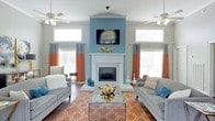 The living room of this single-story space makes an immediate impression through its thoughtfully designed focal point: a corner fireplace framed by a crisp white mantel and surround, set against a bold teal accent wall that extends to the ceiling. A wall-mounted flat-screen television sits above the mantel, maximizing both style and functionality. The teal wall is flanked by large windows on either side, dressed in striking two-tone curtains in burnt orange and gray, which allow natural light to filter into the room while contributing to the space's vibrant color palette. Two ceiling fans are symmetrically positioned overhead, enhancing airflow and adding to the room's balanced, well-planned layout. The living area is furnished with a pair of gray sofas arranged symmetrically around a glass-top coffee table, centered on a bold burnt orange geometric-patterned area rug that anchors the seating arrangement. Decorative accents, including gold sculptural pieces and coordinating throw pillows in teal and neutral tones, add layers of texture and visual interest. The overall ambiance is lively yet cohesive, blending warm and cool tones for a dynamic, inviting atmosphere.
