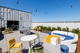 Located at 2121 Wood St in Oakland, CA, this dynamic rooftop terrace is a well-appointed outdoor space designed for gathering, relaxation, and recreation. The terrace is defined by its contemporary industrial aesthetic, with corrugated metal and bold-paneled building facades framing the open-air environment. Generous raised planting beds filled with ornamental grasses and mulched landscaping are distributed throughout the space, lending a natural, park-like quality to the urban setting. String lights crisscross overhead, creating an inviting ambiance suitable for both daytime and evening use. Multiple seating configurations are thoughtfully arranged across the paved terrace, including bold yellow-framed lounge chairs with teal cushions, modular sofas, bar-height tables with chairs, and casual cluster seating — all designed to accommodate a variety of social settings. A Cornilleau ping pong table adds a recreational focal point near the building entrance. At the perimeter, a railed overlook reveals sweeping views of the Oakland skyline and surrounding East Bay landscape beneath open blue skies. The combination of lush greenery, colorful furnishings, and panoramic city views makes this rooftop terrace a distinctive and functional outdoor amenity.