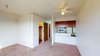 This single-floor residence offers an open and functional layout that combines the living area and kitchen in a practical, connected design. The living space is finished with wall-to-wall carpet in a neutral beige tone, creating a warm and cohesive atmosphere throughout. A ceiling fan with a built-in light fixture is centrally mounted, providing both airflow and ambient lighting to the main area.

The kitchen is partially separated from the living space by a white pass-through counter, which adds visual openness while defining the two zones. Rich warm-toned wood cabinetry is mounted above the kitchen area, offering ample storage and a classic contrast against the white surfaces below. Appliances, including a stove and refrigerator, are visible from the living area through the pass-through, contributing to the kitchen's efficient and compact design.

Toward the back of the unit, two doorways are visible — one leading to what appears to be a closet and another to an additional room, suggesting dedicated storage and private spaces. The neutral wall tones and simple finishes throughout the unit create a clean, understated aesthetic that is easy to personalize.