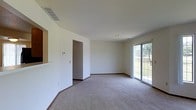 This two-story home offers a clean, functional layout designed to maximize comfort and usability across both levels. The main living area is generously sized, featuring neutral carpet flooring and crisp white walls that create a bright, open atmosphere. A sliding glass door floods the space with natural light while providing direct access to the outdoors, offering views of the surrounding greenery. A ceiling vent ensures efficient climate control throughout the room, complemented by recessed lighting for a streamlined overhead appearance. A pass-through opening in the wall connects the living area to an adjacent space, likely a kitchen, enhancing the sense of openness and facilitating easy interaction between rooms. The interior doorway leads further into the home, suggesting a well-connected floor plan. Neutral-toned baseboards frame the carpeted floors throughout. The overall aesthetic is understated and versatile, with a simple, move-in-ready design that allows for a wide range of furnishing styles. The two-floor layout provides ample opportunity for dedicated living and sleeping zones across the home.