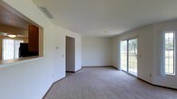 This two-story home offers a clean, functional layout designed to maximize comfort and usability across both levels. The main living area is generously sized, featuring neutral carpet flooring and crisp white walls that create a bright, open atmosphere. A sliding glass door floods the space with natural light while providing direct access to the outdoors, offering views of the surrounding greenery. A ceiling vent ensures efficient climate control throughout the room, complemented by recessed lighting for a streamlined overhead appearance. A pass-through opening in the wall connects the living area to an adjacent space, likely a kitchen, enhancing the sense of openness and facilitating easy interaction between rooms. The interior doorway leads further into the home, suggesting a well-connected floor plan. Neutral-toned baseboards frame the carpeted floors throughout. The overall aesthetic is understated and versatile, with a simple, move-in-ready design that allows for a wide range of furnishing styles. The two-floor layout provides ample opportunity for dedicated living and sleeping zones across the home.