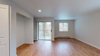 Located at 12456–12498 SE Whitcomb Ave in Milwaukie, Oregon, this single-floor unit offers a thoughtfully designed open layout with consistent hardwood-style flooring throughout. The living area is bright and spacious, benefiting from multiple windows that invite ample natural light and a sliding glass door that opens to an outdoor area with views of surrounding greenery. Baseboard heating units line the walls, ensuring comfortable climate control year-round.

The kitchen is well-appointed with white shaker-style cabinetry extending to the upper and lower walls, light-toned countertops, a stainless steel sink, and a full suite of stainless steel appliances including a gas range, dishwasher, and refrigerator. A peninsula-style counter partially separates the kitchen from the living space, maintaining an open and connected feel.

The bedroom area features a built-in closet with a hanging rod and shelving, providing practical storage. A bathroom is cleanly finished with light-colored walls, wood-style flooring, a vanity with cabinet storage, and a toilet. The overall interior palette of soft whites and neutral grays lends the space a clean, modern ambiance that is both functional and inviting.