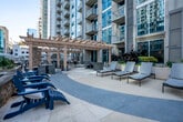 This outdoor amenity deck is situated at podium level within an urban high-rise setting, offering a thoughtfully designed retreat surrounded by the city skyline. The centerpiece of the space is a generously sized swimming pool with blue mosaic tile detailing, flanked by a wide, multi-toned concrete and terracotta-patterned pool deck. Rows of contemporary chaise lounges are positioned along the pool's edges, providing ample space for sunbathing while taking in the surrounding tower views.

Adjacent to the pool area, a substantial pergola structure crafted from warm natural wood defines an outdoor lounge and dining zone. String lights are strung across the pergola's open-beam ceiling, creating an inviting atmosphere for both daytime and evening gatherings. Beneath the pergola, multiple seating arrangements feature dark metal dining tables and chairs, complemented by wicker lounge seating. A built-in stone fireplace serves as a focal architectural element within this covered area, adding warmth and character.

The deck's perimeter is lined with metal railings that open to views of the surrounding downtown streetscape and neighboring high-rise buildings. Potted plants and landscaped planters are integrated throughout, softening the urban hardscape. The overall design balances modern materials with comfortable outdoor living amenities.