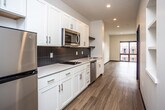 This single-floor unit showcases a contemporary design with warm hardwood-style flooring running throughout the open layout. The galley-style kitchen is well-equipped with stainless steel appliances, including a full-size refrigerator, built-in microwave, dishwasher, and a cooktop. White shaker cabinetry with sleek hardware lines the walls, complemented by light countertops and a bold dark tile backsplash that adds contrast. Recessed lighting illuminates the kitchen and hallway, which leads into a spacious, open living area. The living space is bright and airy, featuring a large floor-to-ceiling window with a dark frame that frames street-level views of the surrounding neighborhood, drawing in generous natural light. A wall-mounted mini-split HVAC unit provides climate control. Built-in shelving along one wall offers convenient storage. The bathroom maintains a clean, minimalist aesthetic with white subway tile surrounding a tub-shower combo, a dark-framed vanity with a white countertop, and large-format tile flooring. Overall, the unit presents a cohesive, modern palette with a functional, open feel.