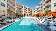 This thoughtfully designed outdoor amenity space is set within a contemporary multi-story residential community, characterized by its warm neutral and yellow-toned exterior siding, expansive balconies, and large grid-pane windows. At the heart of the space is a generously sized swimming pool with crystal-clear turquoise water, flanked on both sides by rows of sleek lounge chairs ideal for sunbathing. A series of vibrant orange-canopied private cabanas line the pool's perimeter, offering shaded retreats with a resort-like atmosphere.

Surrounding the pool, the landscaped grounds feature manicured green spaces, layered plantings, and paved walkways lined with oversized spherical planters bursting with colorful flowers. Intimate seating areas are positioned throughout, furnished with deep-cushioned lounge chairs in navy rope weave, accented with patterned throw pillows and complemented by cheerful yellow and green side tables, creating inviting pockets for relaxation or casual conversation.

A cyber café with floor-to-ceiling glass doors and windows opens directly onto the patio, seamlessly connecting the indoor and outdoor spaces. The overall ambiance is vibrant yet refined, combining modern architectural design with well-curated landscaping to create a welcoming, community-oriented outdoor environment.