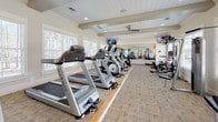 This fitness center is thoughtfully designed to support a comprehensive workout experience within a single, open-plan floor. The space is defined by its attractive architectural details, including exposed ceiling beams and a white shiplap ceiling finish that lend a clean, polished aesthetic. Recessed lighting and ceiling fans maintain a comfortable environment throughout.

Large, shuttered windows line the walls on multiple sides, flooding the interior with natural light and offering views of surrounding wooded scenery. The generous layout accommodates a full row of Matrix-brand cardio equipment, including treadmills, elliptical trainers, and stationary bikes, positioned along a dedicated hardwood-trimmed platform. A separate strength training zone features multi-station cable machines, free weights with a dumbbell rack, and a flat bench, making the space suitable for varied fitness routines.

A large wall-mounted mirror in the strength area visually expands the space while allowing users to monitor their form. Multiple flat-screen televisions are mounted throughout, providing entertainment during workouts. Stability balls and additional accessories are wall-mounted for convenient access. The neutral-toned carpet flooring ties the space together, creating a balanced and inviting fitness environment.
