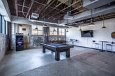 This game room in Memphis, TN, showcases a compelling industrial aesthetic defined by exposed structural ceilings with visible ductwork, original concrete floors, and raw textured concrete walls that lend an authentic, warehouse-inspired character to the space. The open layout provides generous room to move freely between attractions and social areas.

At the center of the space sits a substantial wooden pool table with a dark felt surface, serving as the room's focal point. A wooden ping-pong table occupies an adjacent area, offering additional recreational options. Along one wall, a standalone arcade cabinet provides classic gaming entertainment, while a dartboard mounted on the white-painted wall rounds out the activity offerings.

Multiple high-top tables paired with modern bar stools are distributed throughout the room, creating comfortable spots for socializing and spectating. A wall-mounted flat-screen television adds a media element to the space. Pendant-style industrial lights hang from the ceiling, providing warm, focused illumination throughout.

A row of large steel-framed windows along one wall allows ample natural light to filter in while offering views of the surrounding Memphis neighborhood. Glass-panel partition walls on one side connect the game room to adjacent spaces, maintaining an open, airy feel while defining boundaries within the broader facility.