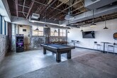 This game room in Memphis, TN, showcases a compelling industrial aesthetic defined by exposed structural ceilings with visible ductwork, original concrete floors, and raw textured concrete walls that lend an authentic, warehouse-inspired character to the space. The open layout provides generous room to move freely between attractions and social areas.

At the center of the space sits a substantial wooden pool table with a dark felt surface, serving as the room's focal point. A wooden ping-pong table occupies an adjacent area, offering additional recreational options. Along one wall, a standalone arcade cabinet provides classic gaming entertainment, while a dartboard mounted on the white-painted wall rounds out the activity offerings.

Multiple high-top tables paired with modern bar stools are distributed throughout the room, creating comfortable spots for socializing and spectating. A wall-mounted flat-screen television adds a media element to the space. Pendant-style industrial lights hang from the ceiling, providing warm, focused illumination throughout.

A row of large steel-framed windows along one wall allows ample natural light to filter in while offering views of the surrounding Memphis neighborhood. Glass-panel partition walls on one side connect the game room to adjacent spaces, maintaining an open, airy feel while defining boundaries within the broader facility.