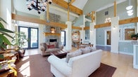 This thoughtfully designed single-floor space makes a striking first impression with its soaring vaulted ceilings and prominent exposed timber beam framework, lending the interior a warm, lodge-inspired character. The expansive great room serves as the heart of the space, anchored by a floor-to-ceiling stacked stone fireplace with a wooden mantel. Comfortable seating arrangements, including plush sofas and classic leather wingback chairs, are positioned around a tufted leather ottoman, creating an inviting gathering area. A dramatic wrought-iron chandelier hangs overhead, complementing the rustic aesthetic. Tall, large-pane windows and glass double doors flood the room with natural light and provide views to the outdoors, while vertical beadboard wall paneling in soft sage green adds a charming, cottage-like texture throughout.

The kitchen is well-equipped with light maple cabinetry, stainless steel appliances, decorative tile backsplash, and light hardwood flooring that flows throughout the space. Ample countertop space and natural light from a nearby window make it both functional and bright. The adjacent dining area features a generous wood table surrounded by upholstered chairs beneath a chandelier, with framed artwork adorning the paneled walls and sheer curtained windows providing a soft, refined ambiance.