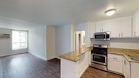 This single-floor residence offers a thoughtfully designed open-concept layout that seamlessly connects the kitchen and living area, creating a bright and functional living space. The kitchen is equipped with stainless steel appliances, including a gas range and over-the-range microwave, complemented by crisp white cabinetry that provides ample storage. A peninsula with granite countertops serves as a natural divider between the kitchen and the living area, offering additional prep space and an opportunity for casual seating. The granite countertops feature warm tones with natural variation, adding a touch of elegance to the space. Rich, dark hardwood-style flooring runs throughout the main living area, providing both durability and a polished aesthetic that ties the space together. The walls are finished in a clean, neutral light gray tone, creating a versatile backdrop that pairs well with a variety of décor styles. A doorway visible from the main area leads to additional rooms, hinting at well-organized interior circulation. The layout and finishes throughout this residence reflect a practical yet refined approach to everyday living.