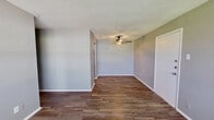 This single-floor space offers a clean, functional layout finished with consistent wood-look flooring throughout, providing a cohesive and warm aesthetic across every room. The main living area is generously proportioned, featuring neutral gray walls, crisp white baseboards, and a ceiling fan with integrated lighting — contributing to a comfortable, well-ventilated environment. A window at the front of the living space invites natural light, offering a view of the surrounding greenery outside.

The kitchen is efficiently designed and equipped with white cabinetry, a speckled light-toned countertop, a double stainless steel sink with a modern brushed-nickel faucet, and a classic white subway tile backsplash that adds a timeless detail to the space. Upper cabinetry provides practical storage, and the layout flows into the adjacent living areas seamlessly.

The bedroom area continues the consistent flooring and neutral palette, with a ceiling fan and access to an interior door, likely leading to a closet or additional storage. The full bathroom is neatly appointed with a toilet, vanity with sink, and a bathtub/shower combination, all finished in white tones that keep the space feeling bright and tidy. Overall, the space presents a straightforward and well-maintained interior with practical finishes throughout.