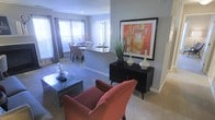This single-floor residence showcases an open-concept layout that connects the living area, dining space, and kitchen in a cohesive, flowing arrangement. The living area is anchored by plush terracotta-hued armchairs and a sleek, low-profile coffee table, creating a warm and inviting focal point. A bold, vibrant abstract artwork in orange and red tones hangs above a mid-century modern console table, which is styled with elegant ceramic vessels and a tall potted plant, adding a sculptural touch to the space.

The kitchen and dining area are visible in the background, featuring a peninsula with a stone or granite countertop that doubles as a casual breakfast bar with seating. Multiple windows dressed with horizontal blinds and soft curtain panels allow natural light to permeate throughout, illuminating the neutral gray walls and wall-to-wall carpeting. The open sightlines between zones create a sense of generous, breathable space. The overall design aesthetic balances contemporary and transitional styles, achieving a polished yet comfortable ambiance suitable for both everyday living and entertaining.