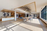 Located at 960 Greene Street in Ann Arbor, Michigan, this sophisticated single-floor space serves as the welcoming lobby and communal lounge for Hoover & Greene. The open-plan layout is generously proportioned, offering a seamless flow between a reception area, a collaborative lounge, and a living room-style gathering space. Upon entry through the glass-paneled automatic doors, polished terrazzo-style flooring leads visitors into a bright, airy interior bathed in natural light from expansive floor-to-ceiling windows that frame views of the surrounding Ann Arbor streetscape. A sleek white marble reception desk anchors the entry zone, accented by warm wood slat ceiling details and striking pendant lighting clusters that add visual drama overhead. The lounge area features a long communal high-top table surrounded by stylish woven chairs, ideal for casual collaboration. A built-in kitchenette with white cabinetry and open shelving provides a functional amenity nearby. The living room zone is highlighted by a white marble-surround fireplace with a wall-mounted display, a bold sculptural shelving unit showcasing curated décor, and textured woven area rugs that ground the warm, contemporary aesthetic. Abstract artwork and carefully selected furnishings complete the polished, design-forward ambiance throughout.