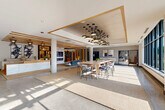 Located at 960 Greene Street in Ann Arbor, Michigan, this sophisticated single-floor space serves as the welcoming lobby and communal lounge for Hoover & Greene. The open-plan layout is generously proportioned, offering a seamless flow between a reception area, a collaborative lounge, and a living room-style gathering space. Upon entry through the glass-paneled automatic doors, polished terrazzo-style flooring leads visitors into a bright, airy interior bathed in natural light from expansive floor-to-ceiling windows that frame views of the surrounding Ann Arbor streetscape. A sleek white marble reception desk anchors the entry zone, accented by warm wood slat ceiling details and striking pendant lighting clusters that add visual drama overhead. The lounge area features a long communal high-top table surrounded by stylish woven chairs, ideal for casual collaboration. A built-in kitchenette with white cabinetry and open shelving provides a functional amenity nearby. The living room zone is highlighted by a white marble-surround fireplace with a wall-mounted display, a bold sculptural shelving unit showcasing curated décor, and textured woven area rugs that ground the warm, contemporary aesthetic. Abstract artwork and carefully selected furnishings complete the polished, design-forward ambiance throughout.