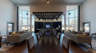 This elegant single-floor community clubhouse is designed to impress, combining modern sophistication with warm, inviting details throughout. The centerpiece of the space is a grand open-concept gathering area anchored by a lengthy dark wood communal dining table surrounded by upholstered chairs, ideal for large group events or community dinners. Overhead, a striking black gridded ceiling feature with pendant lighting defines the dining zone and draws the eye toward the fully equipped kitchen beyond.

The kitchen is outfitted with dark cabinetry, a dramatic dark subway tile backsplash, a spacious quartz-topped island with bar seating, stainless steel appliances, and a built-in television — making it equally suited for casual use or catered gatherings. Flanking the main dining area are comfortable lounge seating arrangements with plush sofas and accent pillows, creating relaxed conversation nooks.

Soaring ceilings lined with floor-to-ceiling windows flood the interior with abundant natural light and provide views of the surrounding exterior. Rich wood-look flooring runs throughout, grounding the contemporary design. Additional highlights include a cozy TV lounge corner, an intimate round table seating area adorned with intricate carved wood wall panels, and a clean elevator corridor with decorative tile accents at the entry.