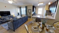 This single-floor residence offers an open-concept layout that integrates the living, dining, and kitchen areas into one cohesive, functional space. The living area is anchored by a plush navy blue sofa and features warm hardwood-style flooring that extends throughout, lending continuity and a sense of spaciousness. Patterned curtains frame the generous windows, allowing ample natural light to fill the space and creating a welcoming ambiance. The dining area accommodates a round table with elegant wood-and-upholstered chairs, styled with tasteful place settings and decorative greenery that adds a touch of life to the room. The kitchen is well-appointed with dark cabinetry, a subway tile backsplash, marble-style countertops, and stainless steel appliances, including a range and refrigerator. A peninsula with bar seating provides additional functionality and serves as a natural dividing point between the kitchen and dining space. Crown molding details along the ceiling add an architectural polish throughout. A hallway visible in the background hints at additional private rooms beyond, completing this thoughtfully designed, light-filled home.