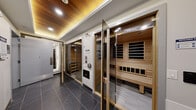 Located at 1792 Pembina Highway in Winnipeg, this thoughtfully designed wellness facility offers a curated suite of amenities within a compact, efficiently arranged single-floor layout. The space features multiple sauna units, including both an infrared sauna and a traditional Finnish sauna, each enclosed with glass-paneled doors and framed in warm natural cedar. The sauna interiors are fitted with tiered wooden benches and dedicated control panels, providing a fully functional and comfortable heat therapy experience.

The main corridor connects each wellness zone seamlessly, finished with large-format dark slate-toned floor tiles that lend a sophisticated, spa-like ambiance throughout. A dedicated shower room offers full wall-to-ceiling dark slate tile cladding, mosaic floor tiles with a central drain, and a glass enclosure — ideal for post-sauna cool-down routines.

A separate relaxation alcove features a solid wood bench set against a rich wood-planked feature wall, offering a serene space to unwind. The ceiling design is a notable architectural highlight, incorporating warm wood paneling with recessed LED lighting that creates an inviting, ambient glow. Control panels and accessibility features are thoughtfully integrated into the corridor walls, ensuring ease of use. The overall design balances natural warmth with modern, clean-lined aesthetics.