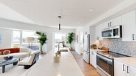 This thoughtfully designed 2-bedroom, 2-bathroom residence spans a single floor and showcases a bright, open-concept layout with clean modern finishes throughout. The entry immediately reveals an expansive living, kitchen, and dining area flooded with natural light from large floor-to-ceiling and oversized windows that frame compelling city views. The kitchen is a standout feature, anchored by a generous waterfall-edge quartz island with bar seating, complemented by white cabinetry, a mosaic tile backsplash, stainless steel appliances, and sleek pendant lighting. The adjacent dining area accommodates a full table and benefits from a corner window with urban skyline views. The living room features a curved architectural wall detail, hardwood flooring, and generous window access to outdoor scenery. The primary bedroom offers closet access and an en-suite bathroom, while the second bedroom features floor-to-ceiling windows with sweeping city views. Both bathrooms are well-appointed; one features a glass-enclosed walk-in shower with decorative mosaic tile and a rain showerhead. A dedicated closet provides additional storage. The residence presents a cohesive, contemporary aesthetic with white walls, wood flooring, and abundant natural light throughout.