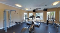 This fitness center is thoughtfully designed to support a well-rounded workout routine. The space features a dark rubberized flooring that provides both durability and cushioning underfoot, ideal for high-impact exercise. Multiple cardio machines, including treadmills and an elliptical trainer, are positioned toward the windowed wall, allowing users to exercise while enjoying views of the outdoor greenery. A multi-station strength training unit is also available for resistance and weight-based workouts, complemented by a weight bench nearby. The warm, neutral-toned walls are finished with classic crown molding, lending a polished, inviting feel to the room. Natural light streams in through several white-framed windows, creating a bright and energizing atmosphere. Overhead, flush-mount light fixtures and a ceiling fan ensure comfortable air circulation and consistent illumination throughout the day. Wall-mounted flat-screen televisions are positioned for easy viewing from the cardio equipment, enhancing the workout experience. The single-floor layout keeps everything accessible and organized, making this fitness center a practical and welcoming space for daily exercise.
