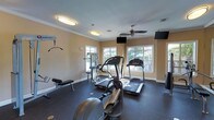 This fitness center is thoughtfully designed to support a well-rounded workout routine. The space features a dark rubberized flooring that provides both durability and cushioning underfoot, ideal for high-impact exercise. Multiple cardio machines, including treadmills and an elliptical trainer, are positioned toward the windowed wall, allowing users to exercise while enjoying views of the outdoor greenery. A multi-station strength training unit is also available for resistance and weight-based workouts, complemented by a weight bench nearby. The warm, neutral-toned walls are finished with classic crown molding, lending a polished, inviting feel to the room. Natural light streams in through several white-framed windows, creating a bright and energizing atmosphere. Overhead, flush-mount light fixtures and a ceiling fan ensure comfortable air circulation and consistent illumination throughout the day. Wall-mounted flat-screen televisions are positioned for easy viewing from the cardio equipment, enhancing the workout experience. The single-floor layout keeps everything accessible and organized, making this fitness center a practical and welcoming space for daily exercise.