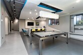 This single-floor amenity space is thoughtfully designed to balance recreation, socializing, and focused work within a contemporary, open layout. The space immediately draws attention with its bold color palette — vivid yellow accent walls contrast against cool grey tones and crisp white surfaces, creating an energetic yet polished atmosphere.

At the heart of the space sits a sleek billiards table, serving as a central social focal point. Adjacent to it, a kitchenette area provides convenient access to refreshments and appliances, supporting both casual gatherings and day-to-day use. A pendant light fixture with a decorative design adds a refined touch overhead.

A dedicated lounge nook offers comfortable seating, complete with a plush sofa accented by teal and patterned throw pillows, a colorful area rug, and a wall-mounted flat-screen television. This area also features a built-in workstation with a monitor and ergonomic chair, making it equally suited for productivity. Vibrant pop-art wall decor adds personality to the space.

Track lighting runs throughout the ceiling, complementing the exposed ductwork in select areas for an industrial-modern aesthetic. Natural light filters in through windows, enhancing the open feel. Restroom facilities are cleanly integrated into the layout, finished with the same grey tones found throughout. The overall ambiance is dynamic, modern, and highly functional.