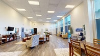Located at 200 E Tenth St in Gilroy, California, this single-story facility is designed to foster community and comfort through thoughtful layout and warm, inviting interiors. The centerpiece of the space is a grand open common area with soaring ceilings and floor-to-ceiling windows along one wall, flooding the room with natural light and offering views of the surrounding outdoors. The area is furnished with multiple comfortable seating arrangements — including sofas, armchairs, and coffee tables — along with dining tables and chairs, creating distinct zones for relaxation and gathering. Durable wood-look flooring and soft area rugs add warmth and texture throughout. A compact but fully equipped kitchen features dark cabinetry, granite-style countertops, a gas range, microwave, coffee maker, and sink, providing functional amenities for daily use. A dedicated workroom offers multiple large tables with seating, a whiteboard, and computer workstations, making it well-suited for structured programs or group activities. Additional areas include a media corner with a wall-mounted television and bar-height seating, as well as tasteful wall art and decorative accents that contribute to the space's welcoming ambiance. The facility is well-lit, accessible, and thoughtfully arranged to serve a variety of needs.