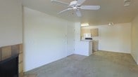 This single-story space features an open-concept layout that connects the living area to the kitchen, creating a functional and cohesive floor plan. The living area is finished with neutral-toned carpeting and smooth, light-colored walls that contribute to a clean and understated aesthetic. A ceiling fan is centrally mounted overhead, providing both air circulation and a subtle decorative element.

A tiled entryway is visible near a white interior door, marking the transition point between the living space and the exterior entrance. The flooring shifts from carpet to tile in this zone, clearly delineating functional areas within the open layout.

The kitchen is positioned at the rear of the main living space, separated by a half-wall or breakfast bar that defines the area while maintaining visual openness. Light wood cabinetry lines the kitchen walls, complemented by white appliances including a range and microwave. Overhead fluorescent lighting illuminates the kitchen work area. The overall design is straightforward and practical, with a neutral color palette throughout that offers a versatile and move-in-ready environment.