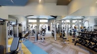 This single-floor fitness center is designed to support a full range of workout routines in a spacious, well-appointed environment. The facility is outfitted with an extensive array of strength-training machines, including cable systems, multi-functional weight stations, and dedicated benches, providing members with everything needed for a complete resistance workout. The open floor plan maximizes usability, with equipment thoughtfully arranged to allow comfortable movement throughout the space. Floor-to-ceiling mirrors line the walls, amplifying the sense of space and allowing users to monitor their form during exercise. The herringbone-patterned flooring adds a modern, polished aesthetic while providing a durable surface suited to heavy use. Elevated windows along the upper portion of the walls usher in generous natural light, contributing to an energizing and welcoming atmosphere. The high, vaulted ceiling enhances the airy feel of the space, preventing any sense of confinement even during peak hours. Soft, neutral tones on the walls complement the contemporary design, while accent colors on the floor add visual interest. Overall, this fitness center strikes a balance between functional design and modern aesthetics, creating an inviting environment for users of all fitness levels.