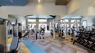 This single-floor fitness center is designed to support a full range of workout routines in a spacious, well-appointed environment. The facility is outfitted with an extensive array of strength-training machines, including cable systems, multi-functional weight stations, and dedicated benches, providing members with everything needed for a complete resistance workout. The open floor plan maximizes usability, with equipment thoughtfully arranged to allow comfortable movement throughout the space. Floor-to-ceiling mirrors line the walls, amplifying the sense of space and allowing users to monitor their form during exercise. The herringbone-patterned flooring adds a modern, polished aesthetic while providing a durable surface suited to heavy use. Elevated windows along the upper portion of the walls usher in generous natural light, contributing to an energizing and welcoming atmosphere. The high, vaulted ceiling enhances the airy feel of the space, preventing any sense of confinement even during peak hours. Soft, neutral tones on the walls complement the contemporary design, while accent colors on the floor add visual interest. Overall, this fitness center strikes a balance between functional design and modern aesthetics, creating an inviting environment for users of all fitness levels.