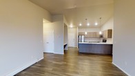 This single-floor space showcases a thoughtfully designed open-concept layout that seamlessly connects the living area to the kitchen. The hardwood-style flooring runs throughout, providing a warm, cohesive aesthetic that ties the space together. Neutral-toned walls complement the overall design, creating a clean and inviting atmosphere.

The kitchen is a standout feature, equipped with stainless steel appliances, including a refrigerator, microwave, and range. Light-toned cabinetry offers ample storage, while a kitchen island with a darker finish provides additional counter space and serves as a natural dividing point between the kitchen and living area. Pendant lighting fixtures hang above the island, adding both functional illumination and a modern design element. Recessed lighting further brightens the kitchen zone.

The entry area is defined by a paneled door and includes a recessed nook, likely suitable for storage or seating. A closet with traditional paneled doors is also visible, offering practical storage solutions. The open sightlines between spaces enhance the sense of roominess, making the layout feel both functional and welcoming.
