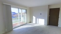 This single-floor space is designed with simplicity and functionality in mind. The main living area is finished with neutral wall-to-wall carpeting and crisp white walls that create a clean, understated backdrop. A standout feature is the large sliding glass door, which floods the room with abundant natural light and provides direct access to an outdoor balcony enclosed by a glass railing. The balcony overlooks a landscaped exterior setting with greenery and neighboring structures visible in the distance, offering a pleasant outdoor connection. Sunlight streams generously across the carpeted floor throughout the day, enhancing the airy feel of the interior. The smooth white ceiling and uncluttered layout contribute to an open, versatile atmosphere well-suited for a variety of furnishing arrangements. A wall-mounted panel is visible near the doorway, indicating built-in utility features. Overall, the space presents a bright, move-in-ready environment with a functional design that balances indoor comfort with easy outdoor access.