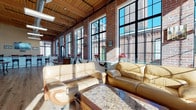 Located at 607 Norwich Avenue in Norwich, CT, this single-floor loft space masterfully blends historic industrial character with modern finishes. The defining architectural feature is the expansive wall of floor-to-ceiling steel-framed windows that line the space, allowing abundant natural light to pour in while offering striking views of the surrounding brick buildings. Original exposed brick walls and warm wood-beamed ceilings lend an authentic industrial aesthetic throughout.

The open main area is anchored by a comfortable lounge seating arrangement featuring tan leather sofas set around a granite-topped coffee table, flowing seamlessly into a dining area with elegant marble-topped tables and wrought iron chairs — all set atop wide-plank hardwood flooring that runs throughout.

A kitchenette area features honey-toned wood cabinetry, granite countertops, and stainless steel appliances, complemented by natural light from adjacent large windows. A separate service bar area offers generous granite-topped cabinetry, a sink, and a wall-mounted display screen, making it well-suited for entertaining or event use.

The overall ambiance is warm and inviting, combining the raw charm of a historic mill building with polished contemporary touches, creating a versatile and visually compelling space.