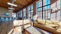 Located at 607 Norwich Avenue in Norwich, CT, this single-floor loft space masterfully blends historic industrial character with modern finishes. The defining architectural feature is the expansive wall of floor-to-ceiling steel-framed windows that line the space, allowing abundant natural light to pour in while offering striking views of the surrounding brick buildings. Original exposed brick walls and warm wood-beamed ceilings lend an authentic industrial aesthetic throughout.

The open main area is anchored by a comfortable lounge seating arrangement featuring tan leather sofas set around a granite-topped coffee table, flowing seamlessly into a dining area with elegant marble-topped tables and wrought iron chairs — all set atop wide-plank hardwood flooring that runs throughout.

A kitchenette area features honey-toned wood cabinetry, granite countertops, and stainless steel appliances, complemented by natural light from adjacent large windows. A separate service bar area offers generous granite-topped cabinetry, a sink, and a wall-mounted display screen, making it well-suited for entertaining or event use.

The overall ambiance is warm and inviting, combining the raw charm of a historic mill building with polished contemporary touches, creating a versatile and visually compelling space.