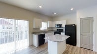 This single-story space showcases a well-appointed kitchen as its centerpiece. The kitchen is designed with clean white cabinetry that lines the walls in an efficient L-shaped layout, providing ample storage throughout. Granite countertops with a subtle grey-and-white pattern run along the perimeter and are echoed on the central island, which offers additional workspace and a natural gathering point. Black appliances, including a refrigerator, electric range, built-in microwave, and dishwasher, create a bold contrast against the white cabinetry. The kitchen island adds functional versatility to the layout. Light hardwood-style flooring flows throughout, contributing to a bright and cohesive aesthetic. Recessed lighting in the ceiling ensures consistent illumination, while large windows and a sliding glass door invite abundant natural light into the space, enhancing its open and airy feel. The neutral wall tones and textured ceiling complement the overall modern yet understated design. The layout is practical and efficiently organized, making the kitchen both visually appealing and highly functional for everyday use.