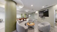 Located at 26 Belden Ave in Norwalk, Connecticut, this single-floor amenity space offers a thoughtfully designed communal environment that blends leisure, socializing, and fitness. The open layout is anchored by polished terrazzo flooring and a cohesive color palette of sage green, warm grey, and crisp white throughout.

The main lounge area features a pool table and an air hockey table, making it a central hub for recreation. Curved, contemporary lounge chairs and rounded sofas are arranged around a wooden coffee table — complete with a chess set — creating an inviting social setting. A wall-mounted flat-screen TV is positioned for comfortable viewing from the seating area.

Architectural interest is provided by a sculptural curved ceiling detail and a distinctive fluted green column, adding depth and character to the space. Glass-paneled walls and doors offer visual connectivity to an adjacent fitness center, which is fully equipped with cardio and strength-training equipment.

A separate dining nook features black tables paired with wishbone-style chairs set against a bold fluted green accent wall, with natural light streaming in through tall windows that overlook the surrounding grounds. Recessed lighting throughout ensures the entire space is well-lit and welcoming at any time of day.